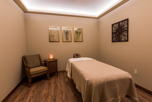 A room with a bed, a chair, a small table with a lamp, and three framed pictures on the wall.