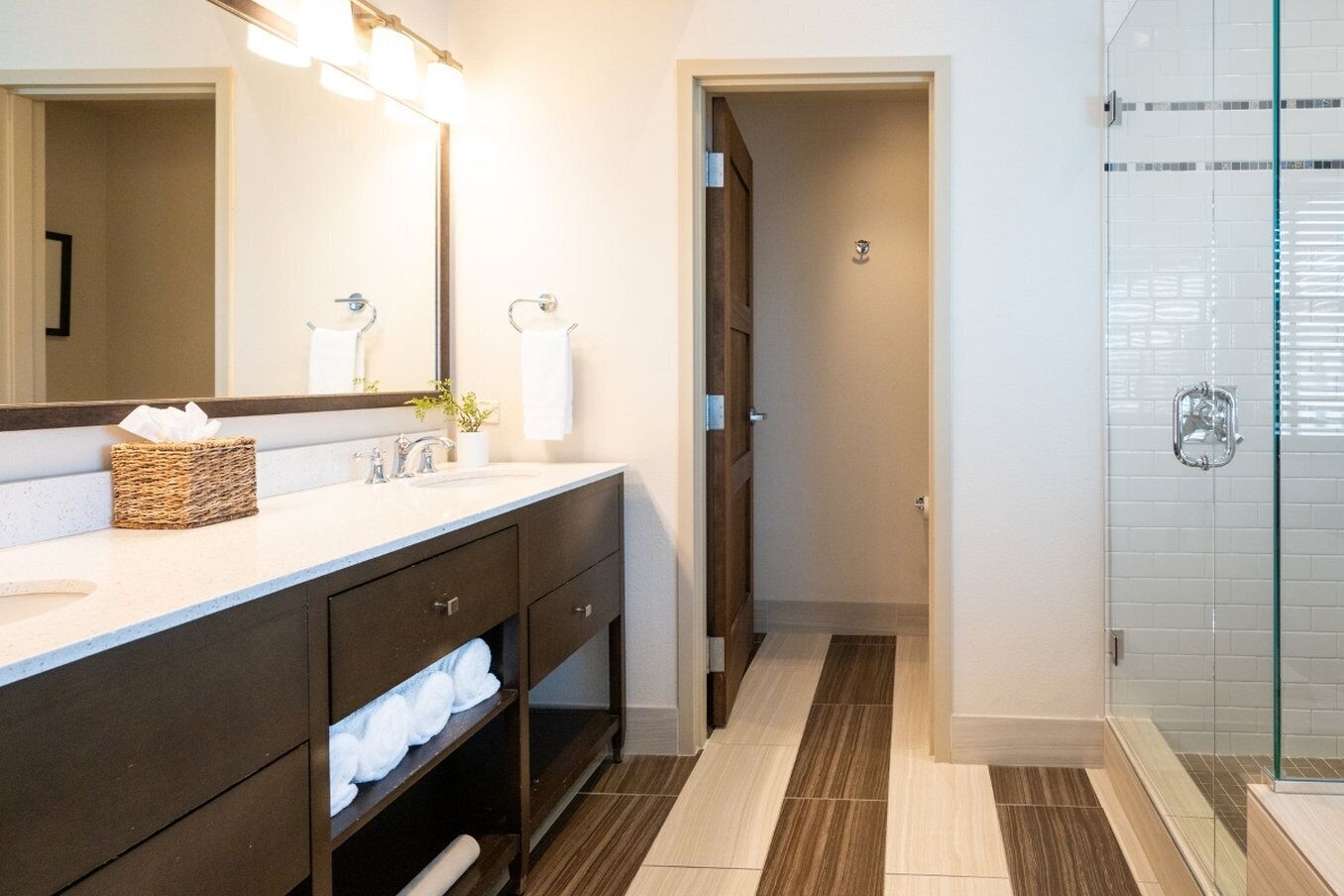 Un baño moderno con un espejo grande, tocador de madera oscura, encimera blanca y cabina de ducha de vidrio.