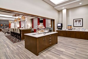 A modern hotel lobby with a central island featuring a breakfast bar, seating area, and a television. - Hampton Inn & Suites by Hilton Grande Prairie (Grande Prairie)