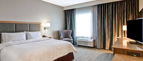 Quarto de hotel com uma cama grande, uma escrivaninha com televisão de tela plana, uma cadeira e uma janela com cortinas.