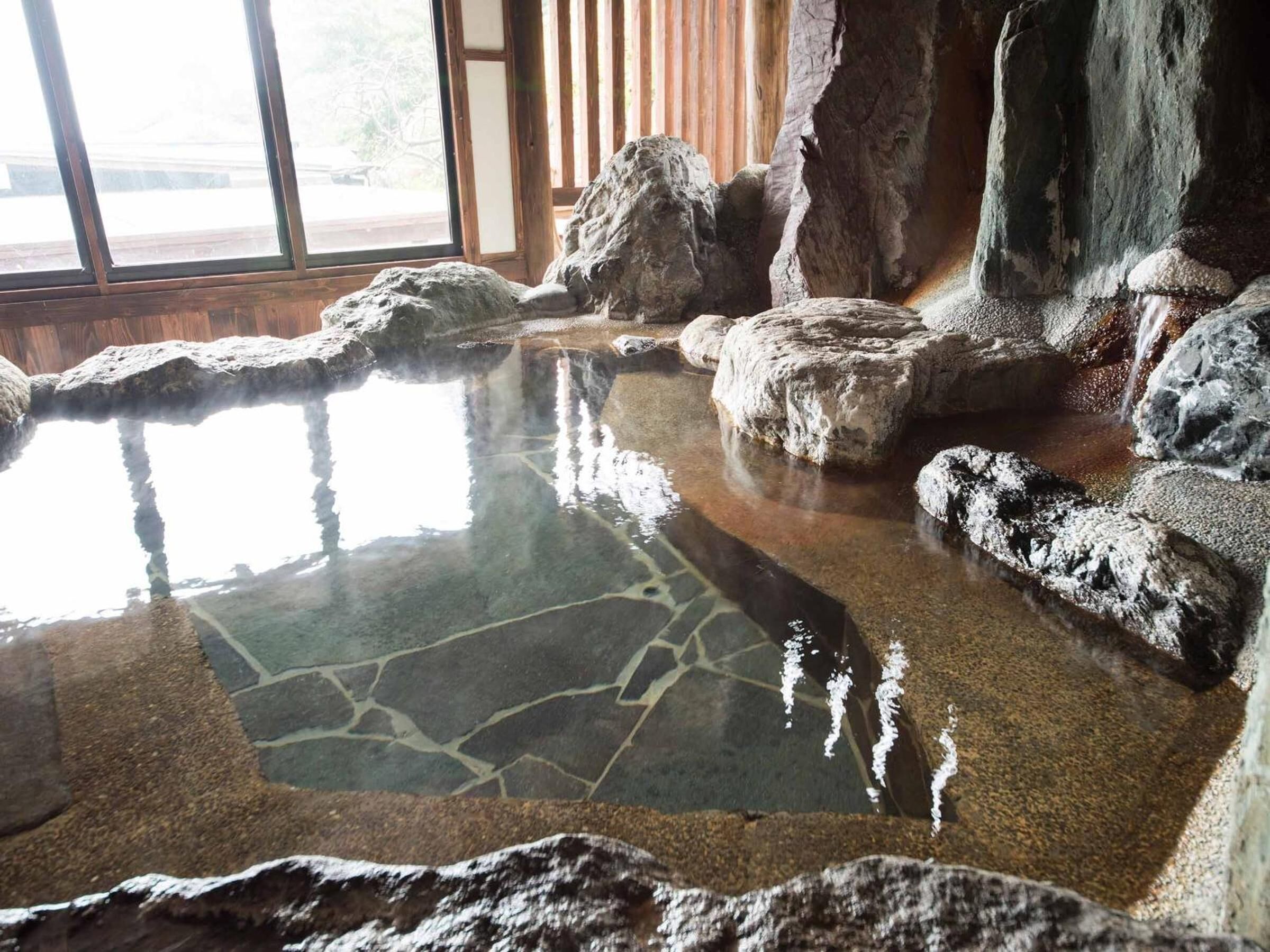 A hot spring area with a stone pool surrounded by rocks, large windows, and a view of the outdoors.