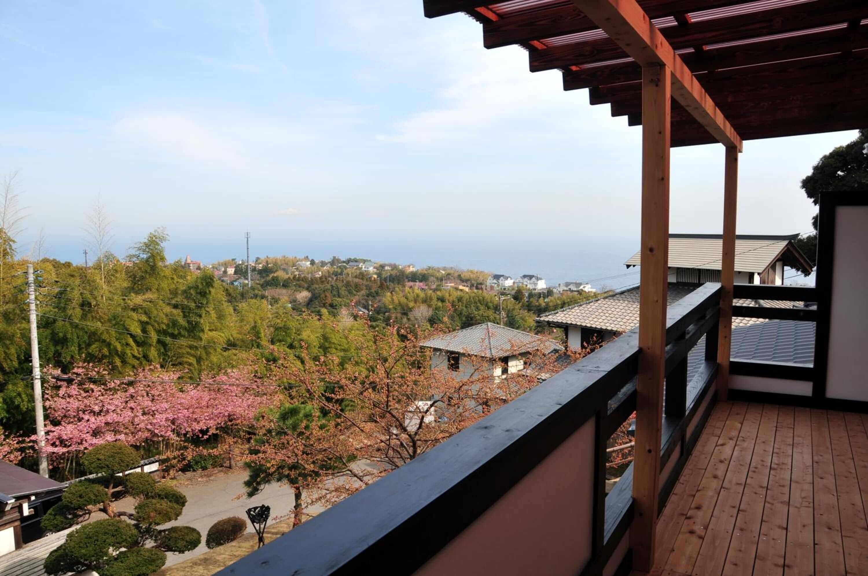 Un balcón de madera con vista a una zona residencial, flores de cerezo y una masa de agua distante.
