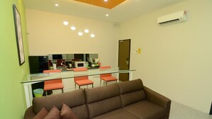 Una sala moderna con un sofá marrón, una mesa de comedor de vidrio con sillas naranjas, una zona de cocina con microondas y un equipo de aire acondicionado en la pared.