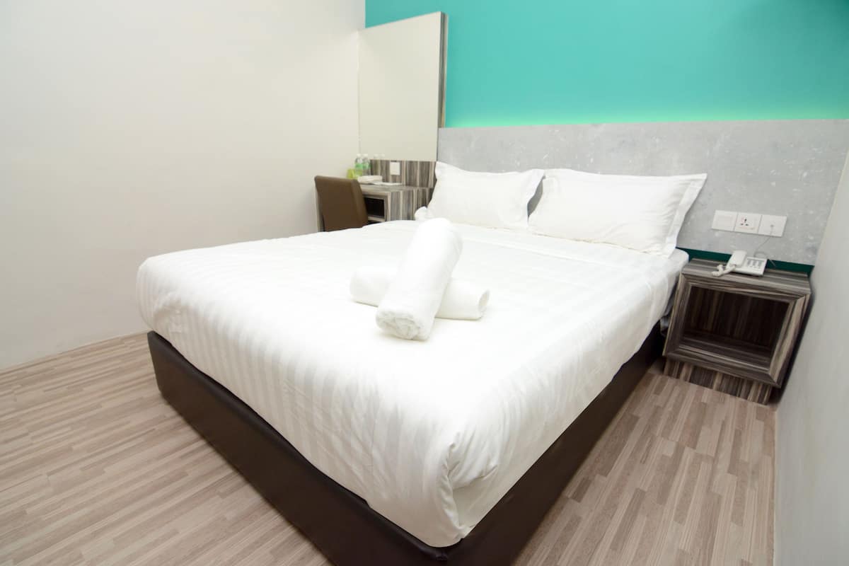 Una cama bien hecha con sábanas blancas y una toalla enrollada, una mesita de noche con un teléfono, y una silla en una habitación de hotel moderna.