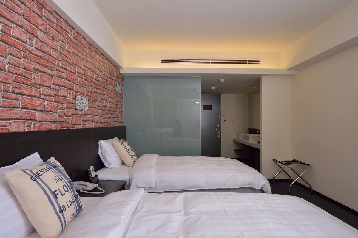 A hotel room with two beds, a brick wall, a glass partition, and a small bathroom area.