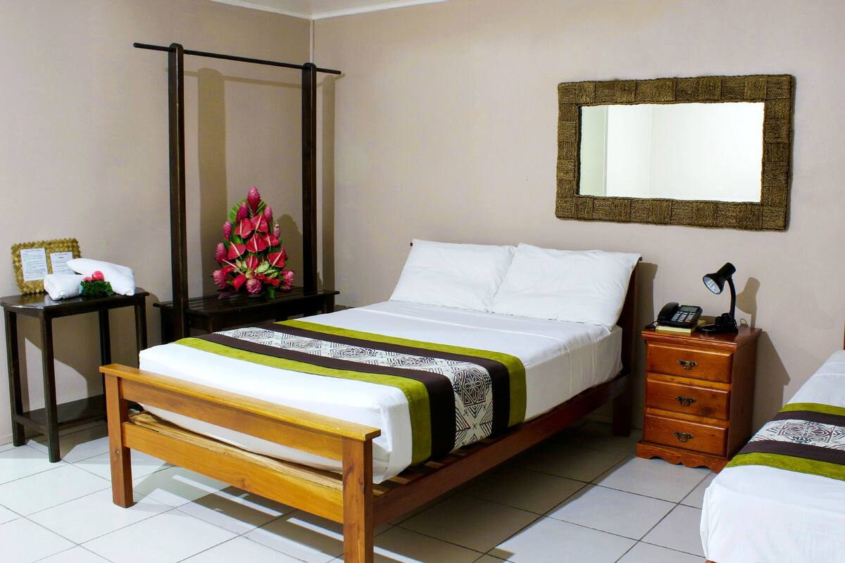Una habitación de hotel con una cama de madera, una mesita de noche con lámpara, un espejo y un arreglo floral decorativo.