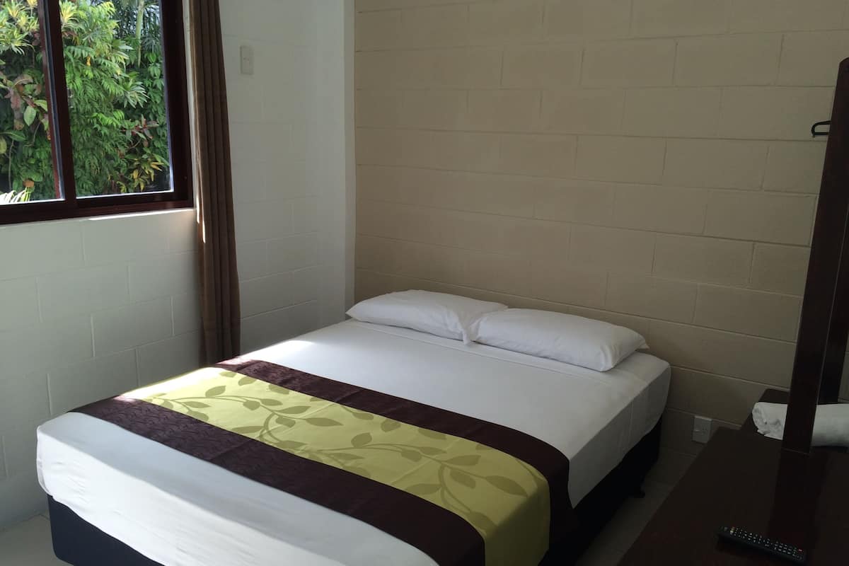 Una cama individual con colchón blanco y edredón marrón y amarillo en una habitación con paredes de ladrillo blanco y una ventana con vistas a la vegetación.