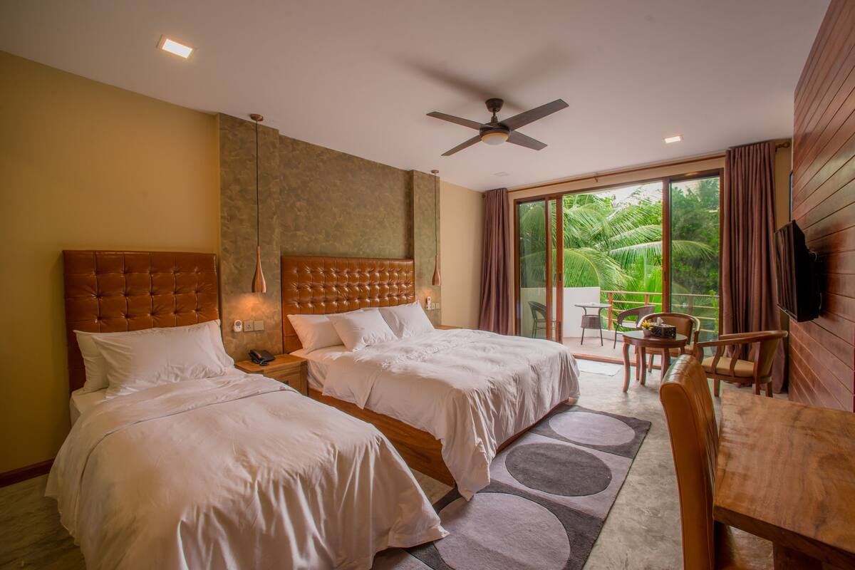 A hotel room with two beds, a ceiling fan, a balcony with a table and chairs, and a TV mounted on the wall.
