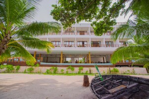 A multi-story hotel with a balcony on the second floor, surrounded by palm trees and a sandy beach.