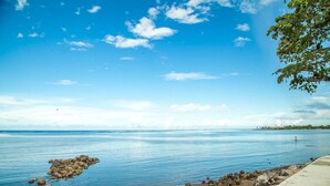 A calm beach with clear blue water, a rocky outcrop, and a concrete walkway along the shore. - Ulalei Lodge (Apia)