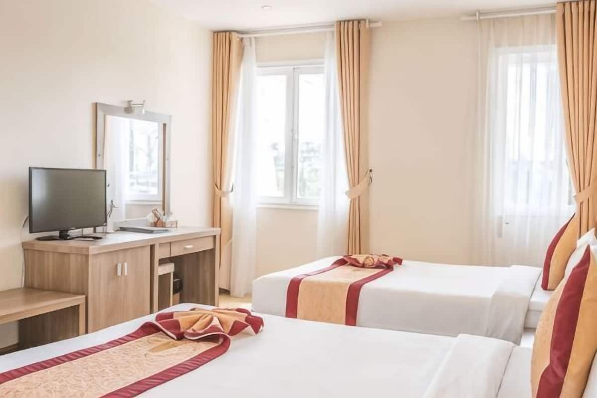 A hotel room with two beds, a desk with a TV, and large windows with curtains.