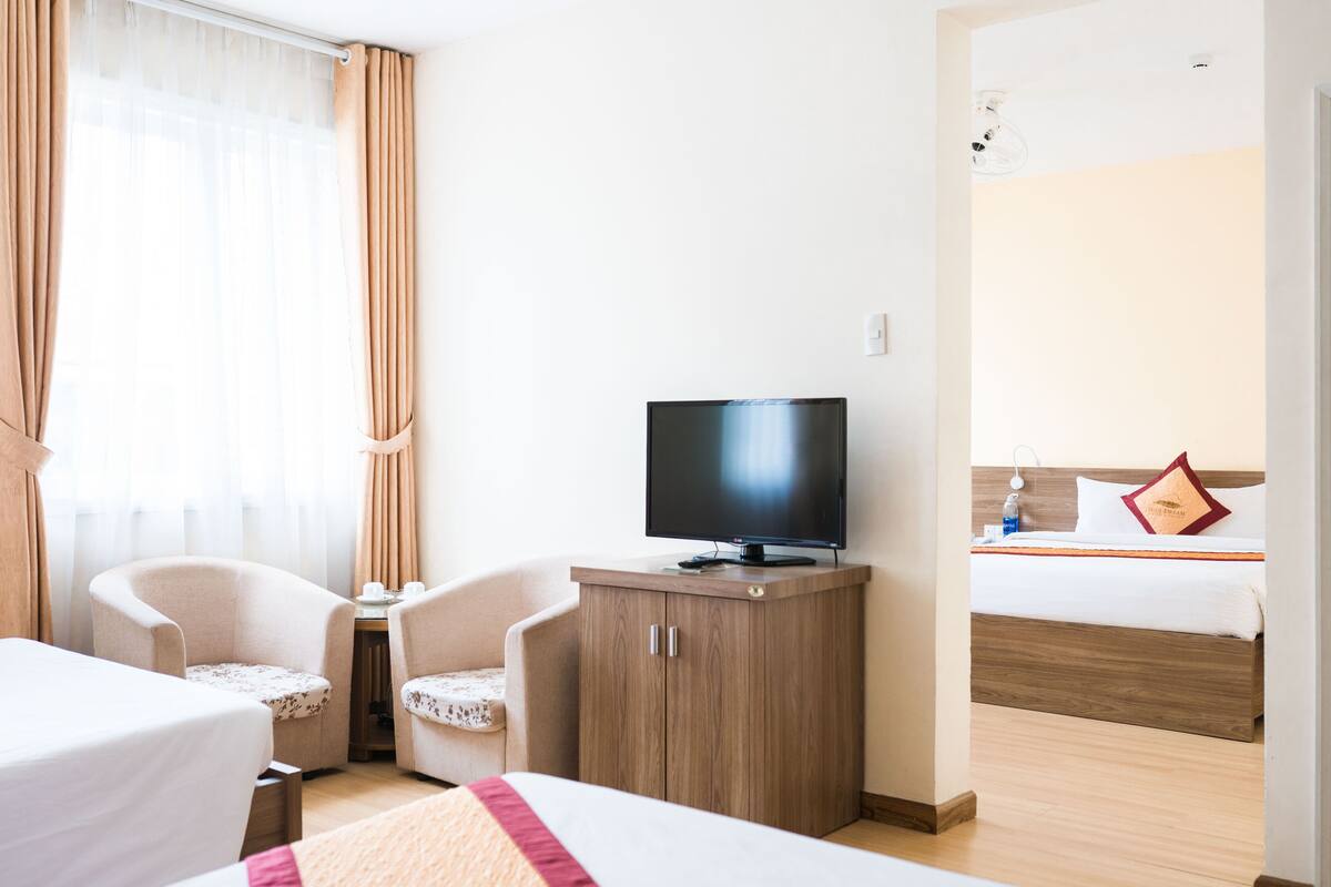 A hotel room with a bed, a TV on a wooden cabinet, two armchairs, a small table, and a window with curtains.