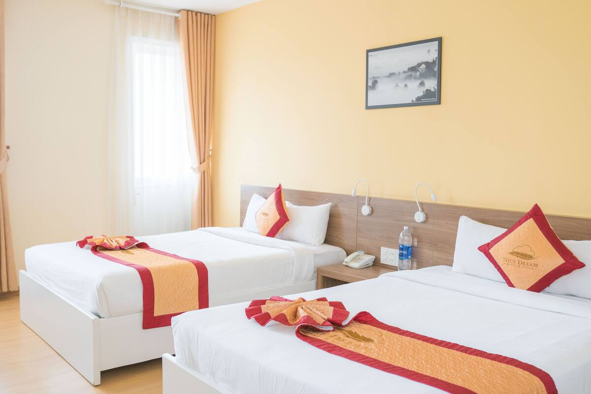 Two single beds with white linens and red accents, a bedside table with a phone and water bottle, and a painting on the wall.