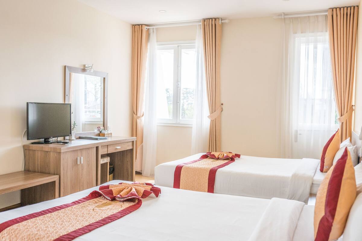 A hotel room with two beds, a TV, a desk, and a window with curtains.