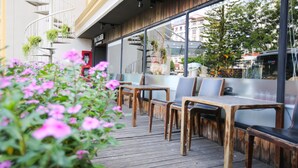 Un área de estar al aire libre con mesas y sillas de madera, una escalera en espiral con plantas y vistas a la calle a través de amplios ventanales.
