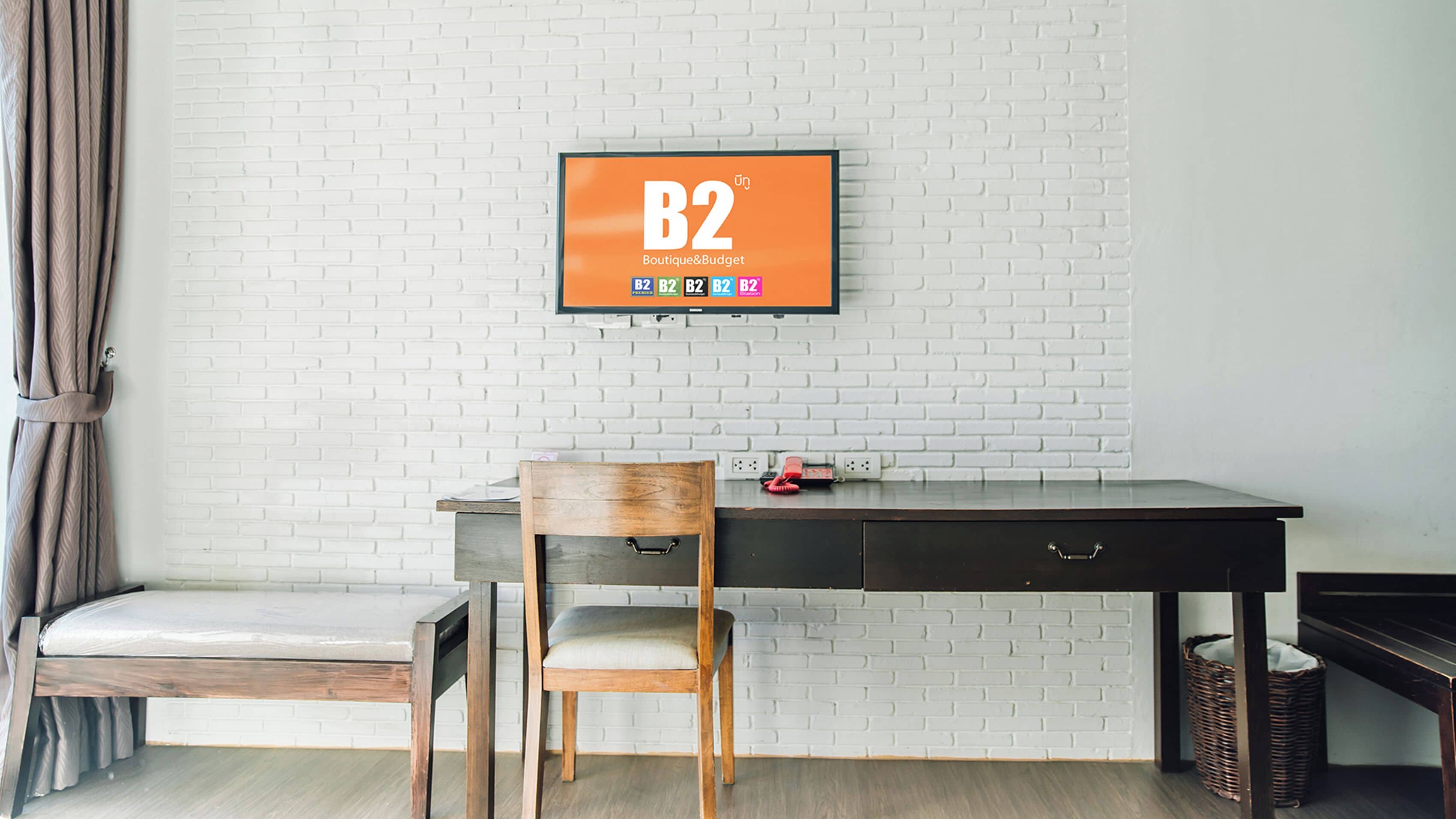A room with a white brick wall, a wooden desk, a chair, a small table, and a television displaying 'B2 Boutique & Budget'.