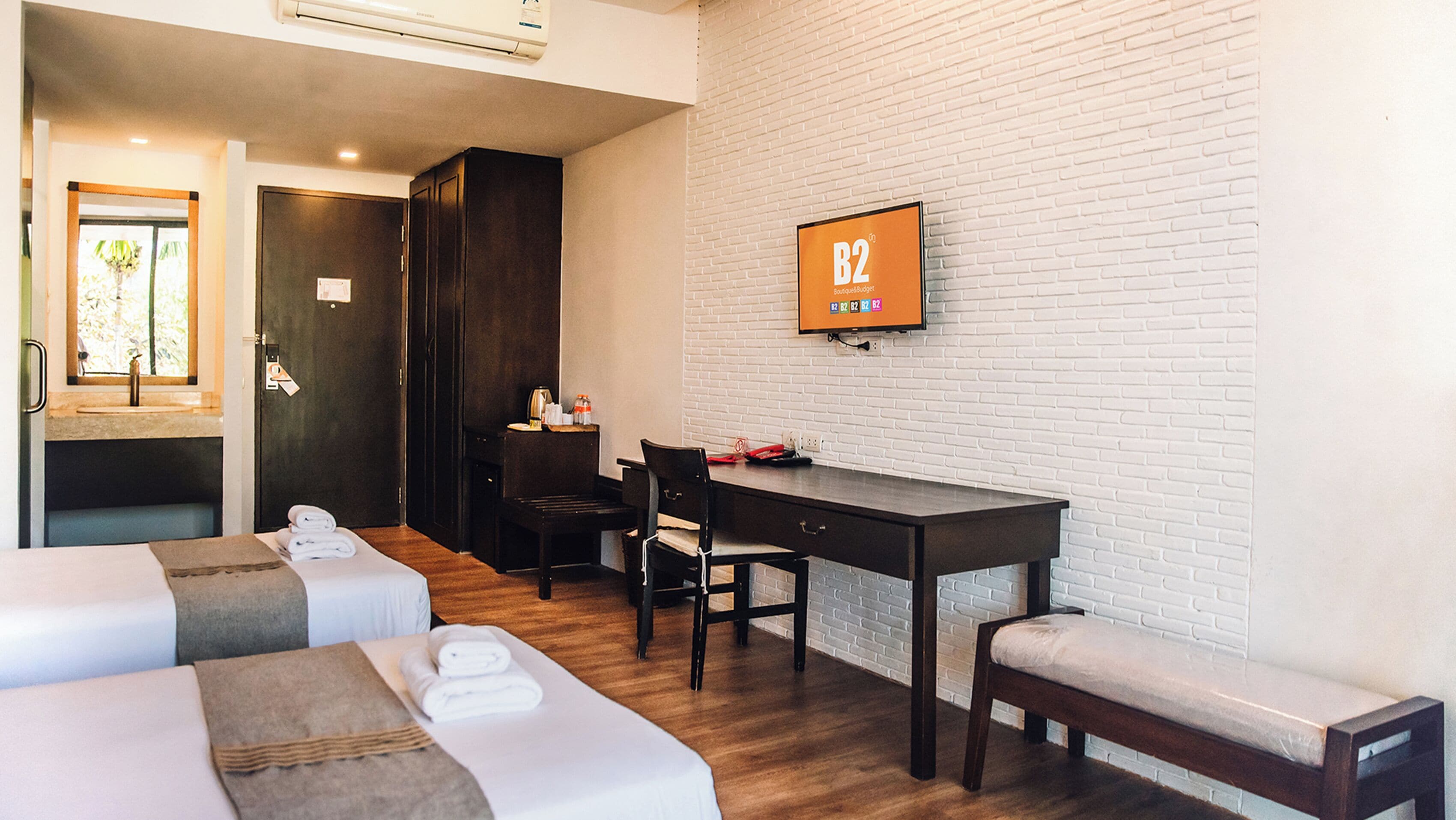 A hotel room with two beds, a desk, a chair, a TV, and a brick wall.
