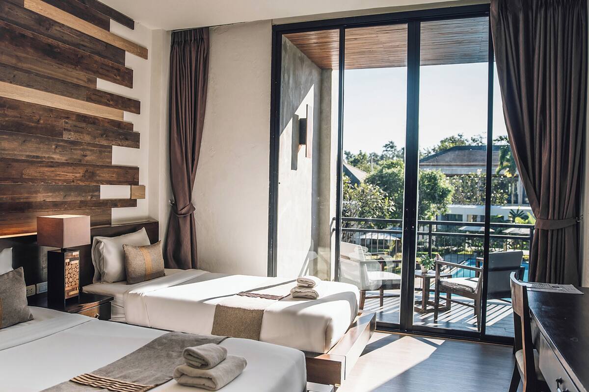 A modern hotel room with two beds, a wooden accent wall, a balcony with a pool view, and a desk.
