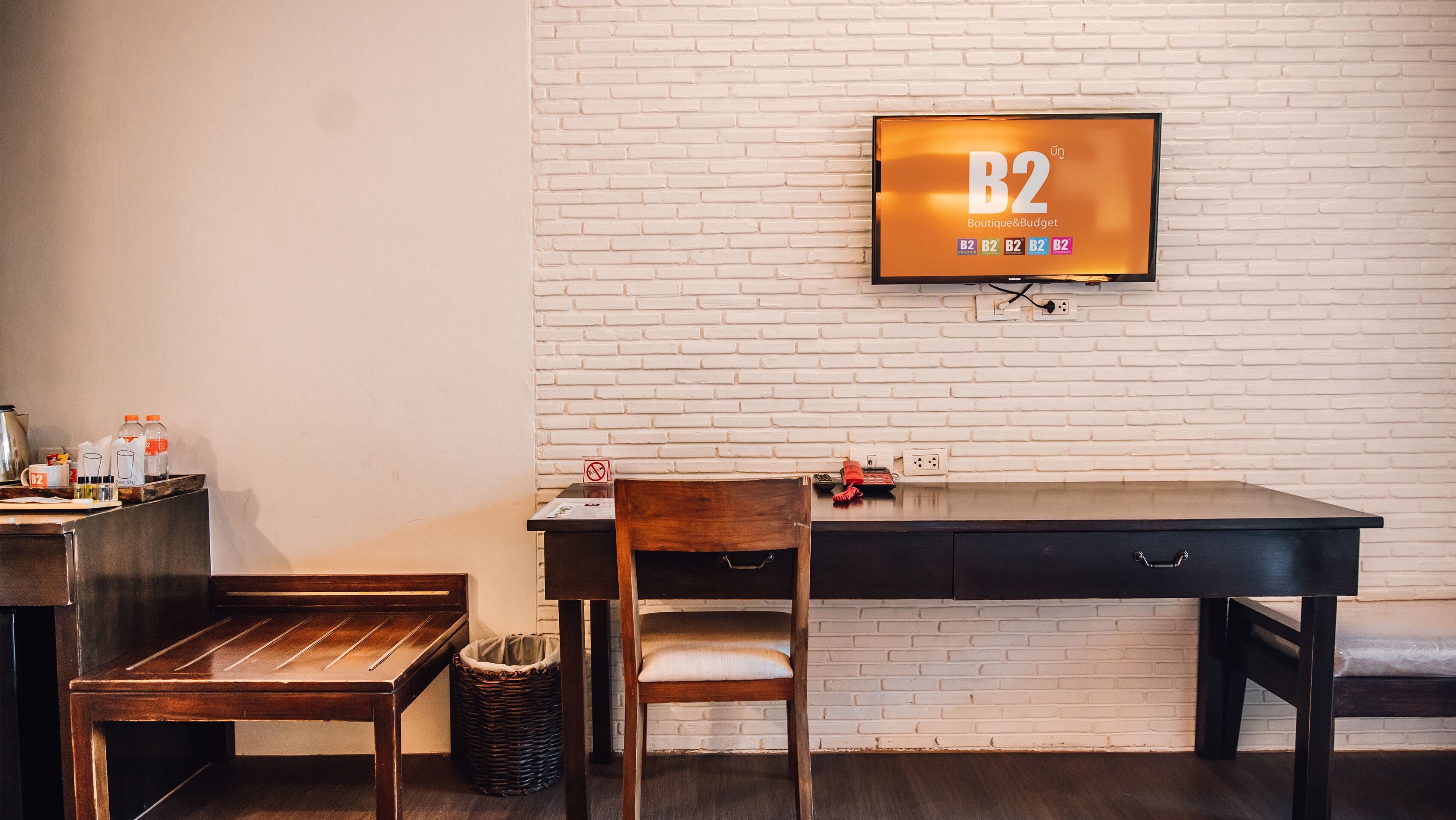 A modern room with a white brick wall, a wooden desk, a chair, a small table, and a television displaying 'B2 Boutique & Budget'.