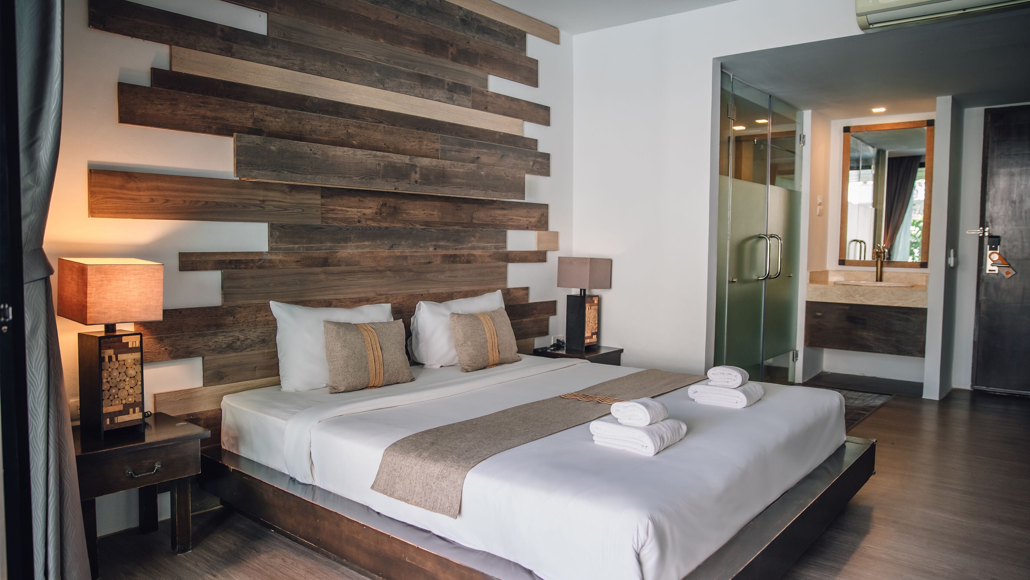 A modern hotel room with a large bed, wooden wall paneling, two bedside lamps, and a bathroom visible through an open door.