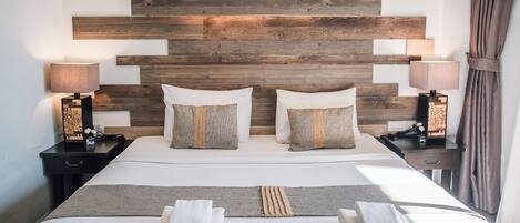Habitación de hotel con cabecera de madera, dos mesitas de noche con lámparas, cama con sábanas blancas y dos almohadas, y un par de toallas dobladas sobre la cama.