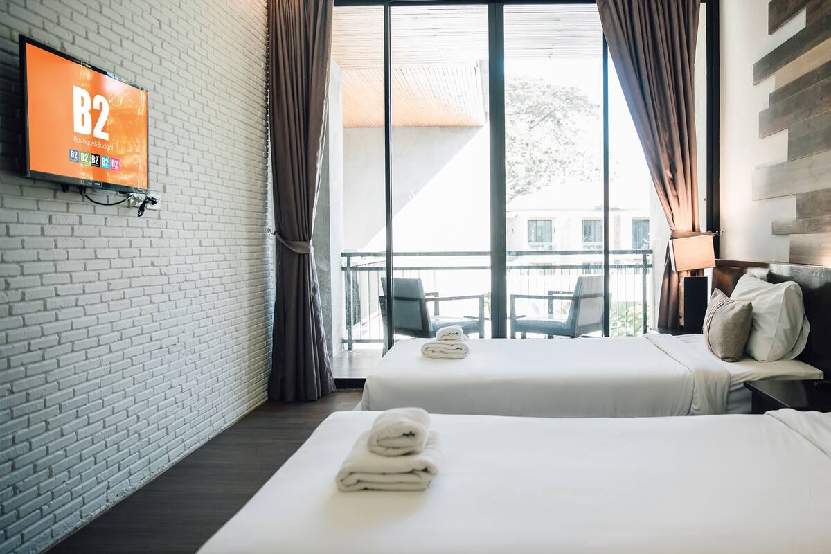 A hotel room with two beds, a TV on the wall, and a balcony with chairs.