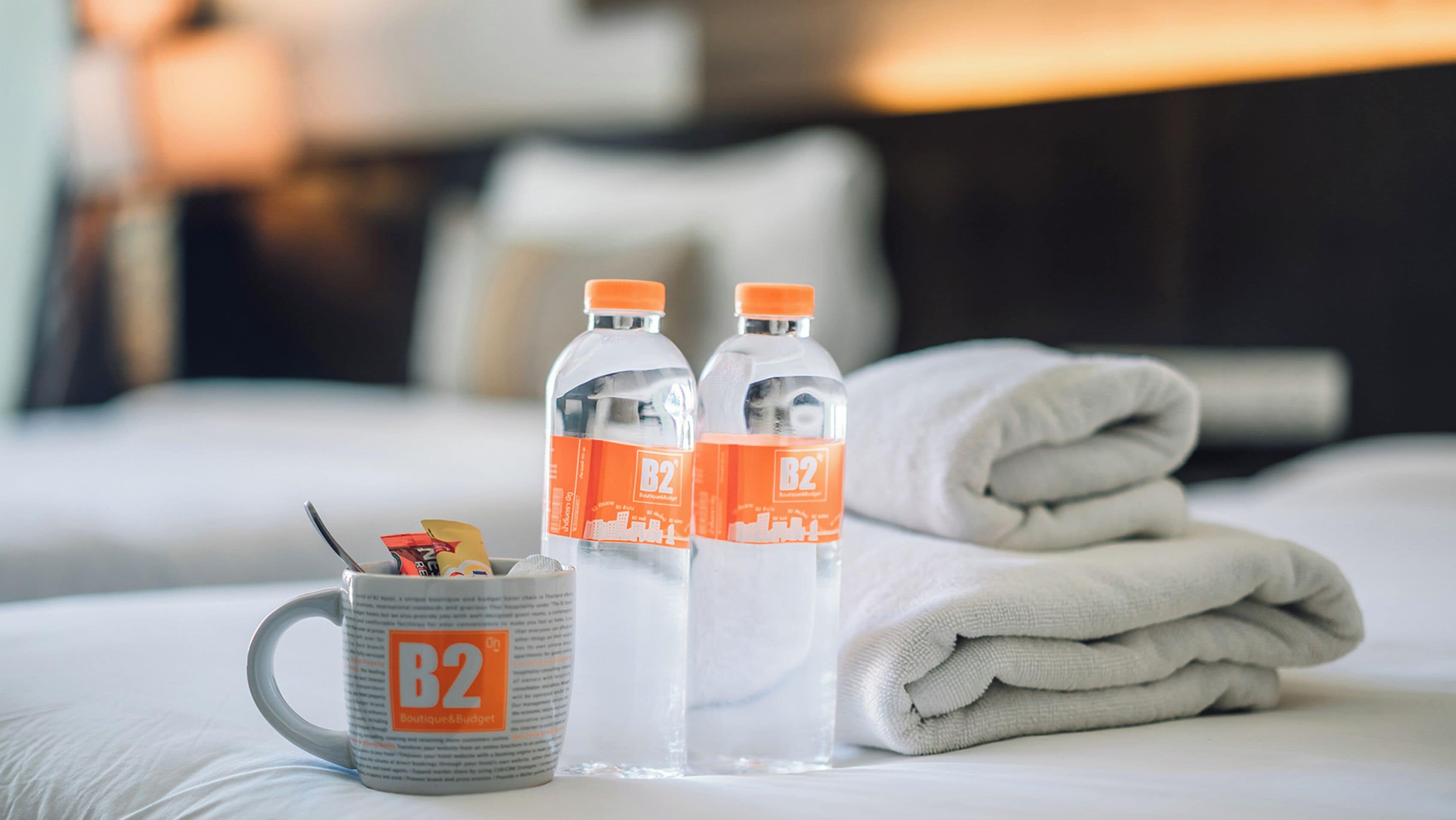Habitación de hotel con una cama bien hecha, dos botellas de agua, una taza y una toalla doblada.
