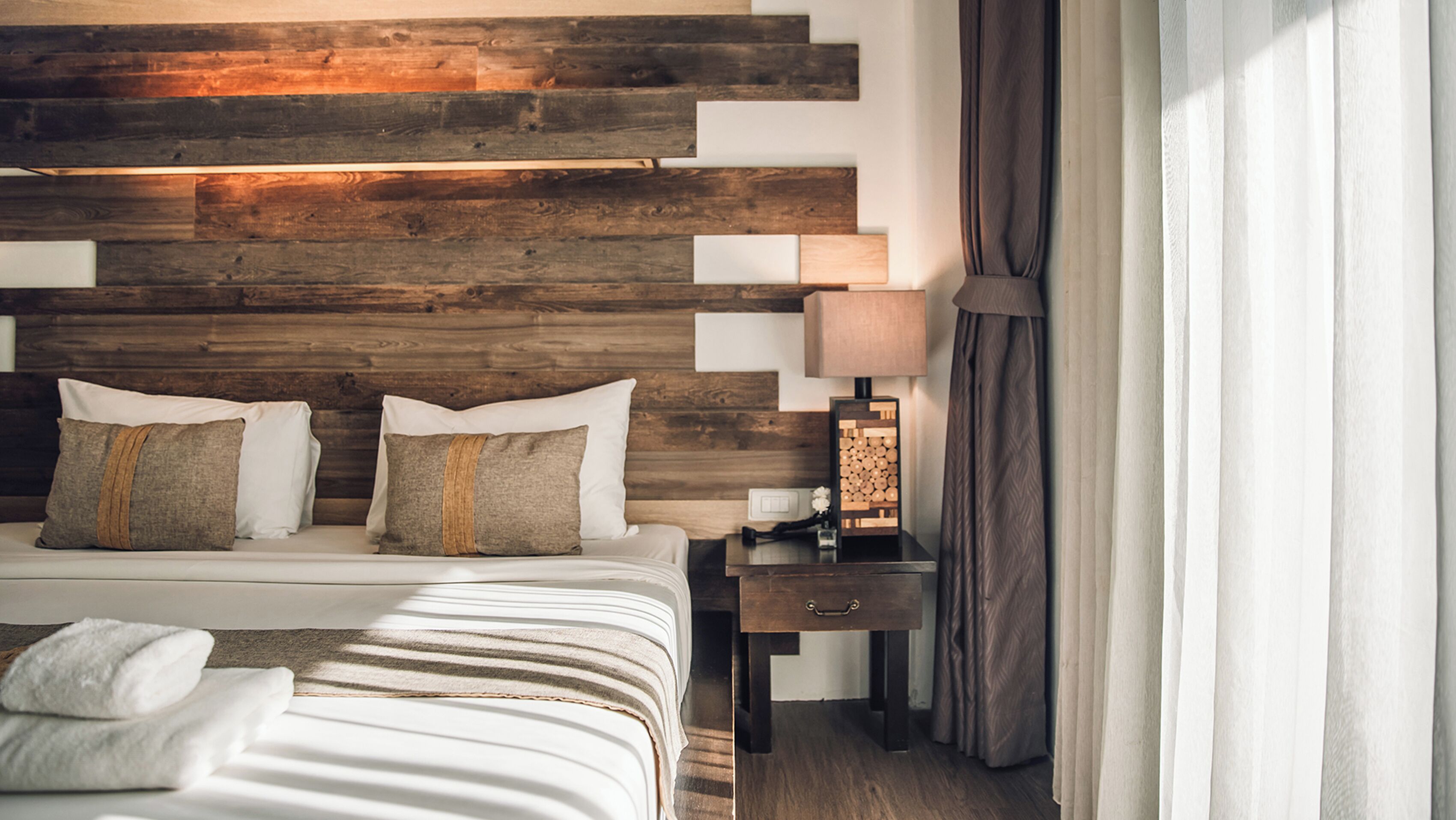 A hotel room with a wooden headboard, a bed with white linens, a bedside table with a lamp, and a window with curtains.