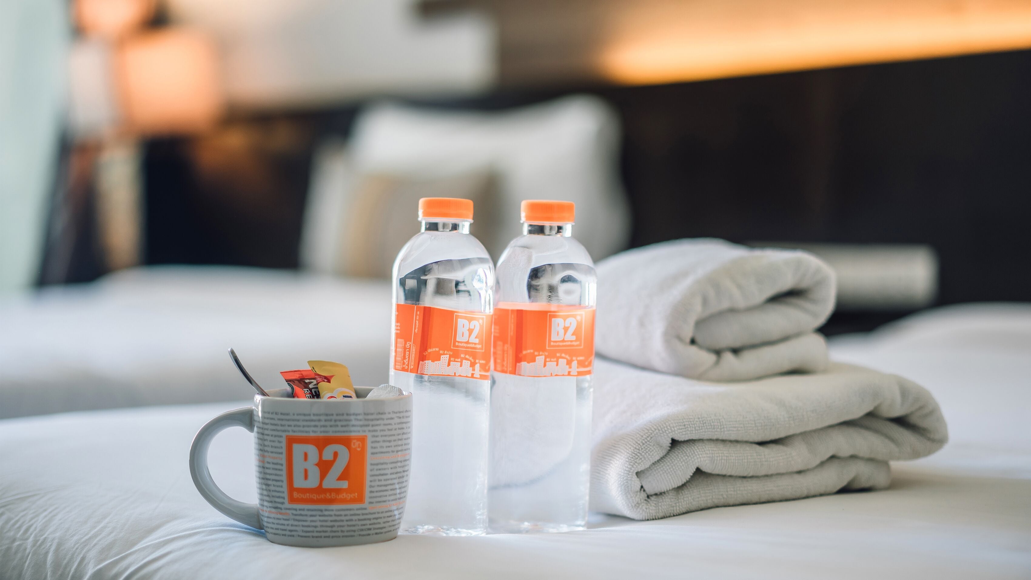 A hotel room with a neatly made bed, two B2 branded water bottles, a mug, and a folded towel on the bed.