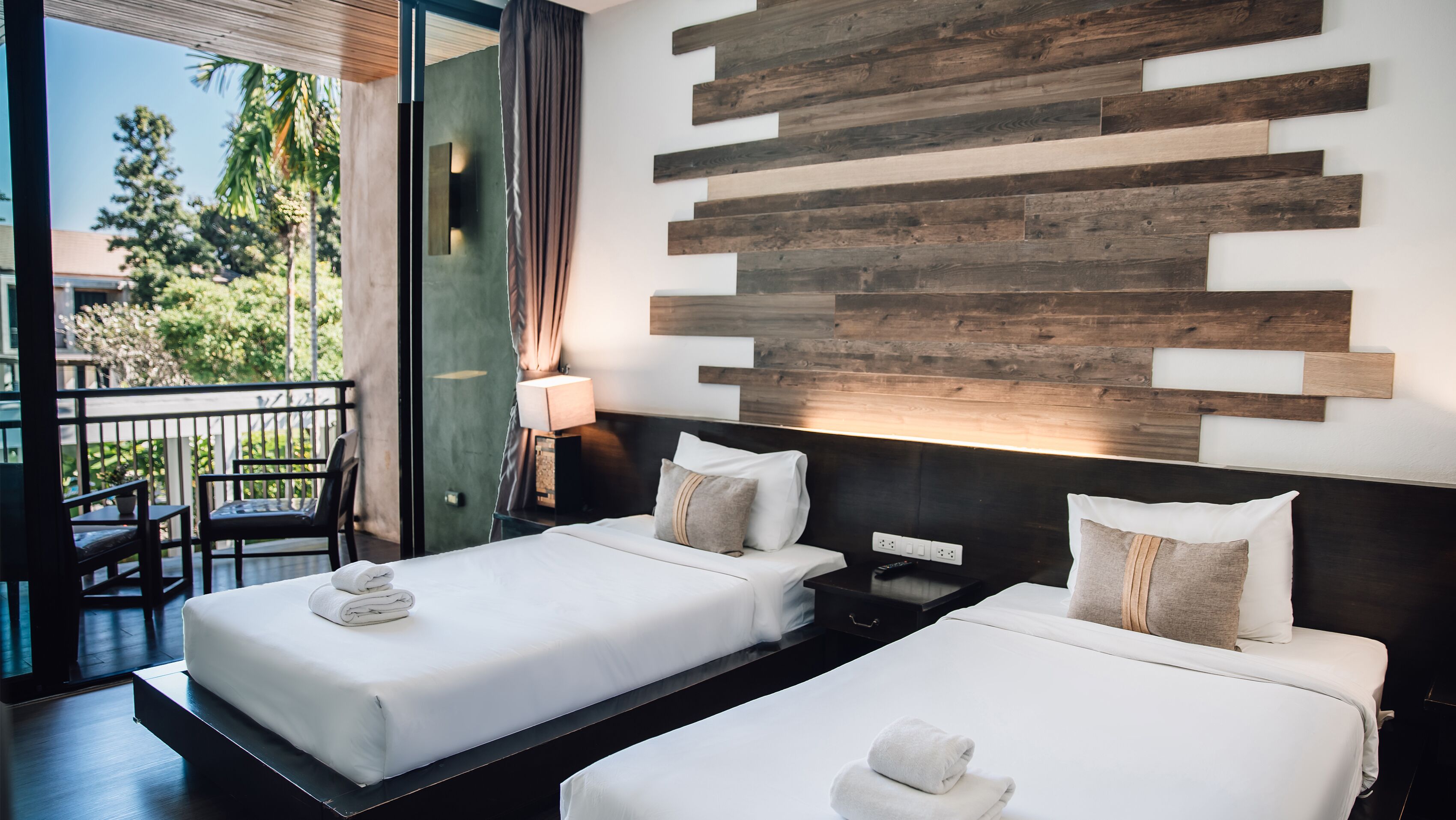 A hotel room with two beds, a wooden accent wall, a balcony with a view of greenery, and a modern design.