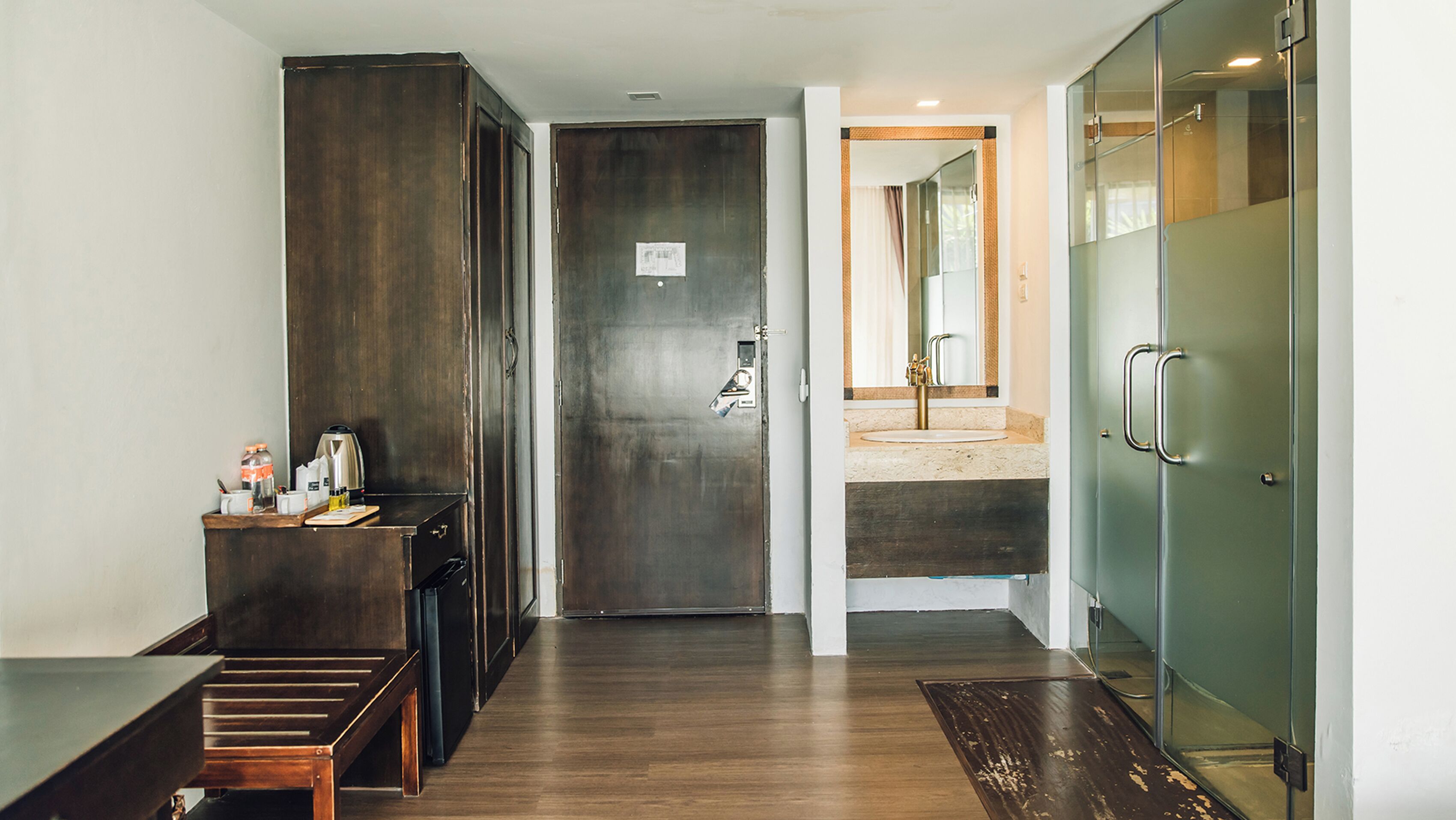 Pasillo de hotel con un banco de madera, un minibar, una puerta y un baño con cabina de ducha de vidrio.