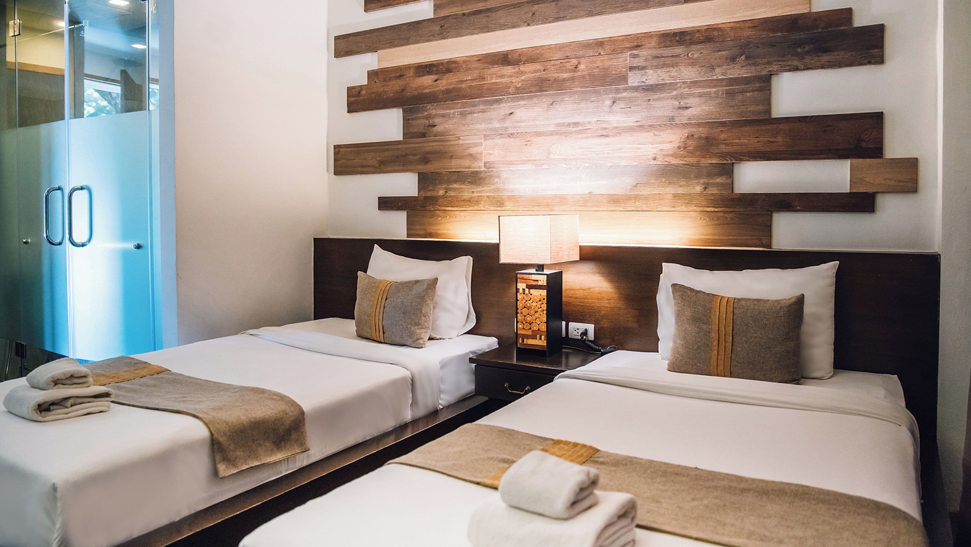 Une chambre d’hôtel avec deux lits, une tête de lit en bois, une table de chevet avec une lampe et une porte en verre donnant sur la salle de bain.