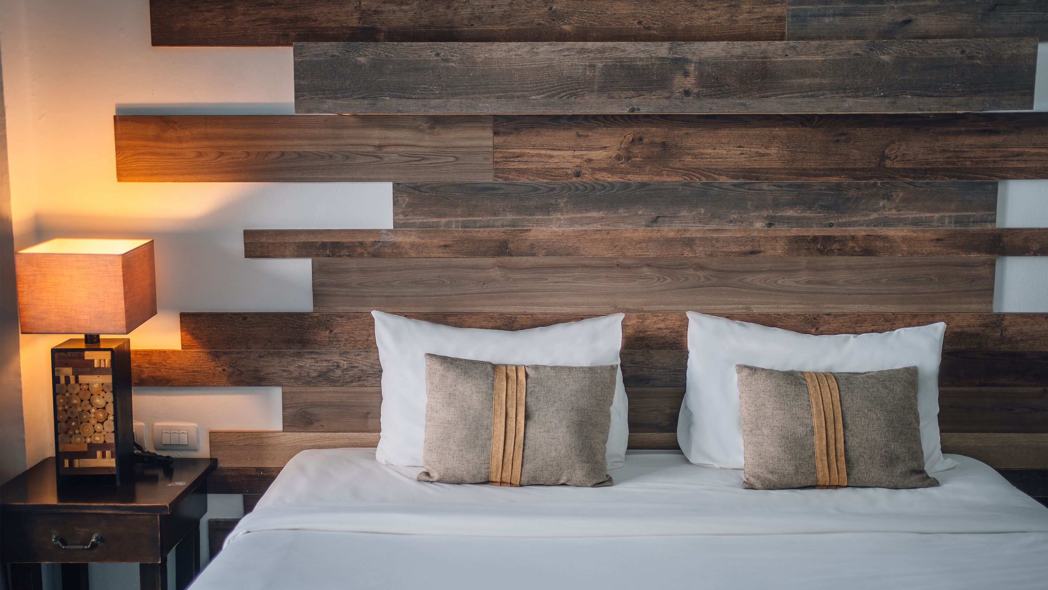 Un dormitorio con cabecera de madera, dos almohadas, una mesita de noche con lámpara y un tocador.