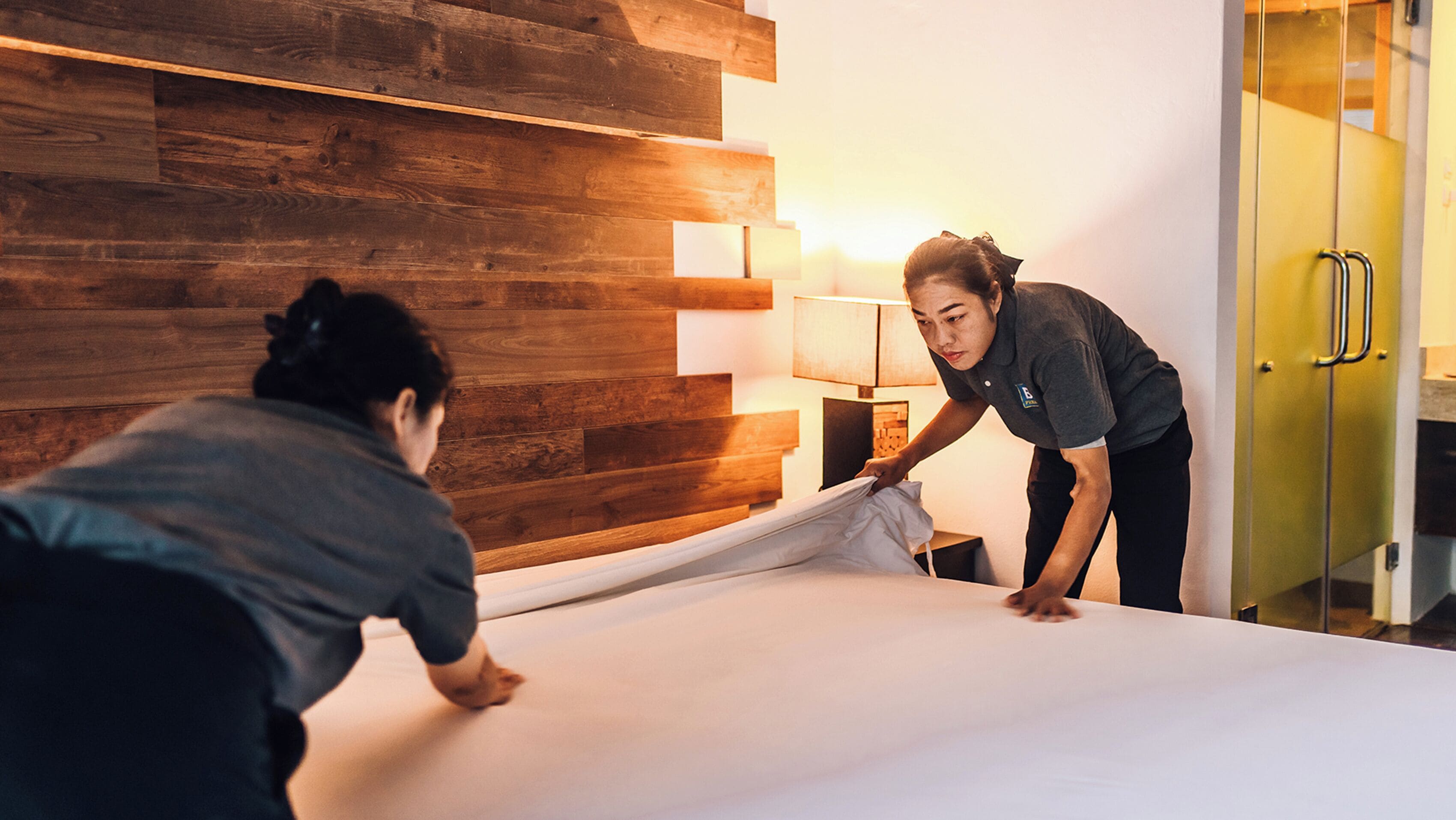 A hotel room with a wooden wall panel, a bed with white linens, and a bedside table with a lamp.