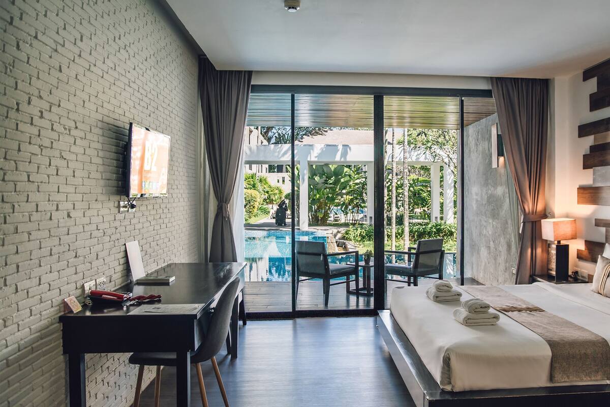 A hotel room with a bed, a desk, a chair, a small table, and a view of a pool and greenery outside.