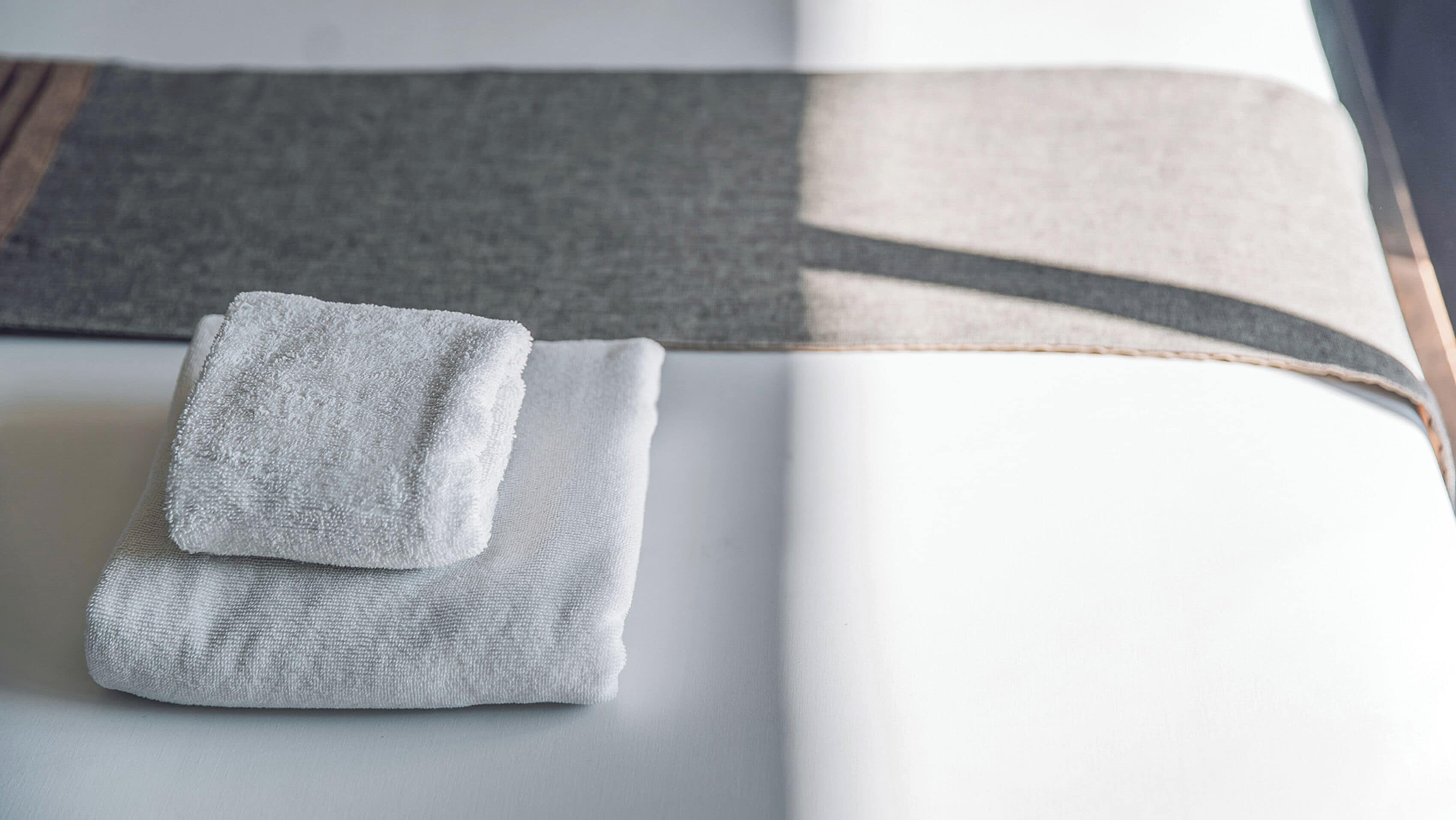 A neatly folded white towel on a bed with a grey and white striped blanket.