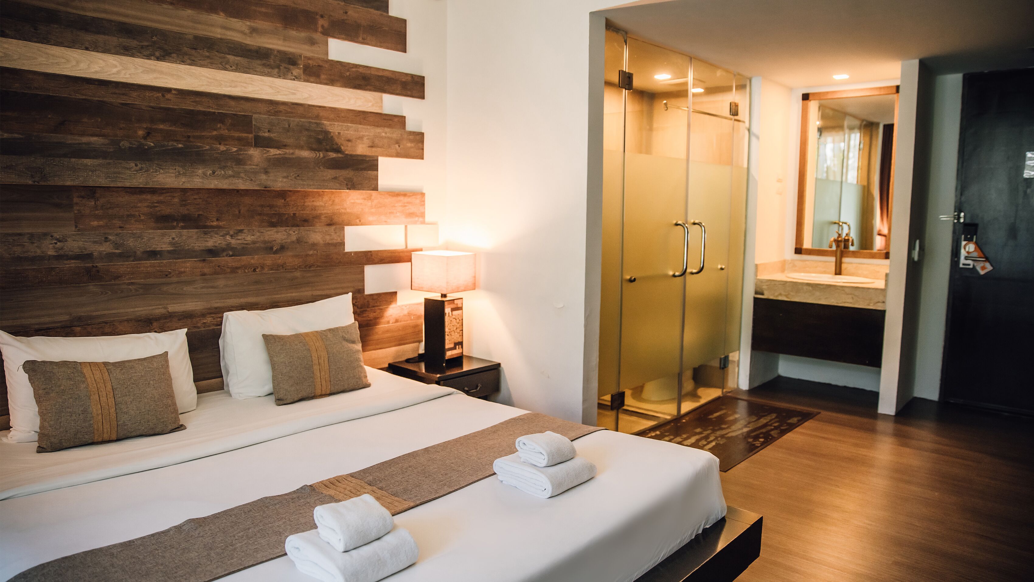 A hotel room with a wooden headboard, a bed with white linens, a nightstand with a lamp, a glass-enclosed shower, and a bathroom with a mirror and sink.