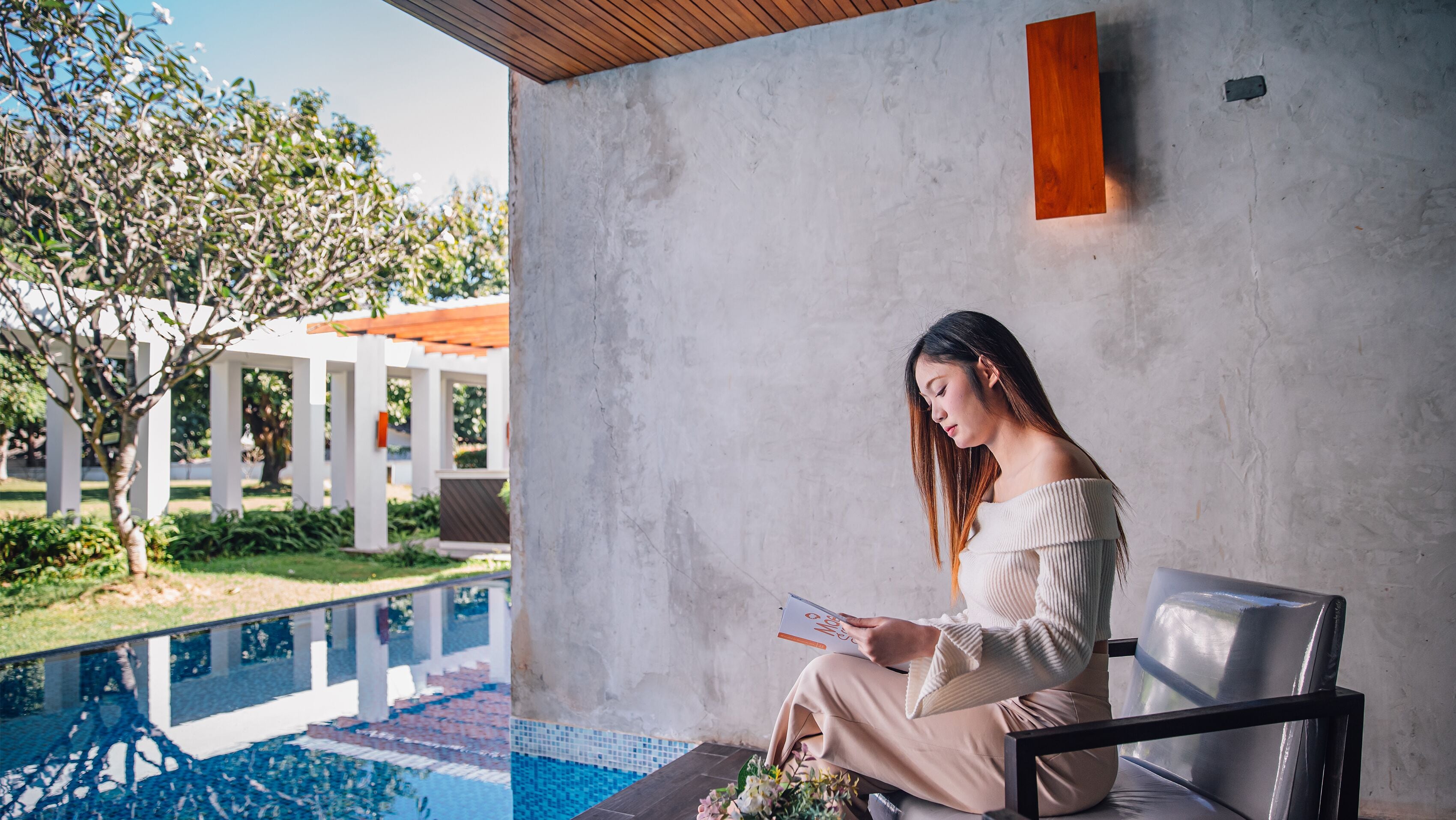 A person sitting by a poolside with a book, in a modern outdoor setting with a concrete wall and a wooden ceiling.