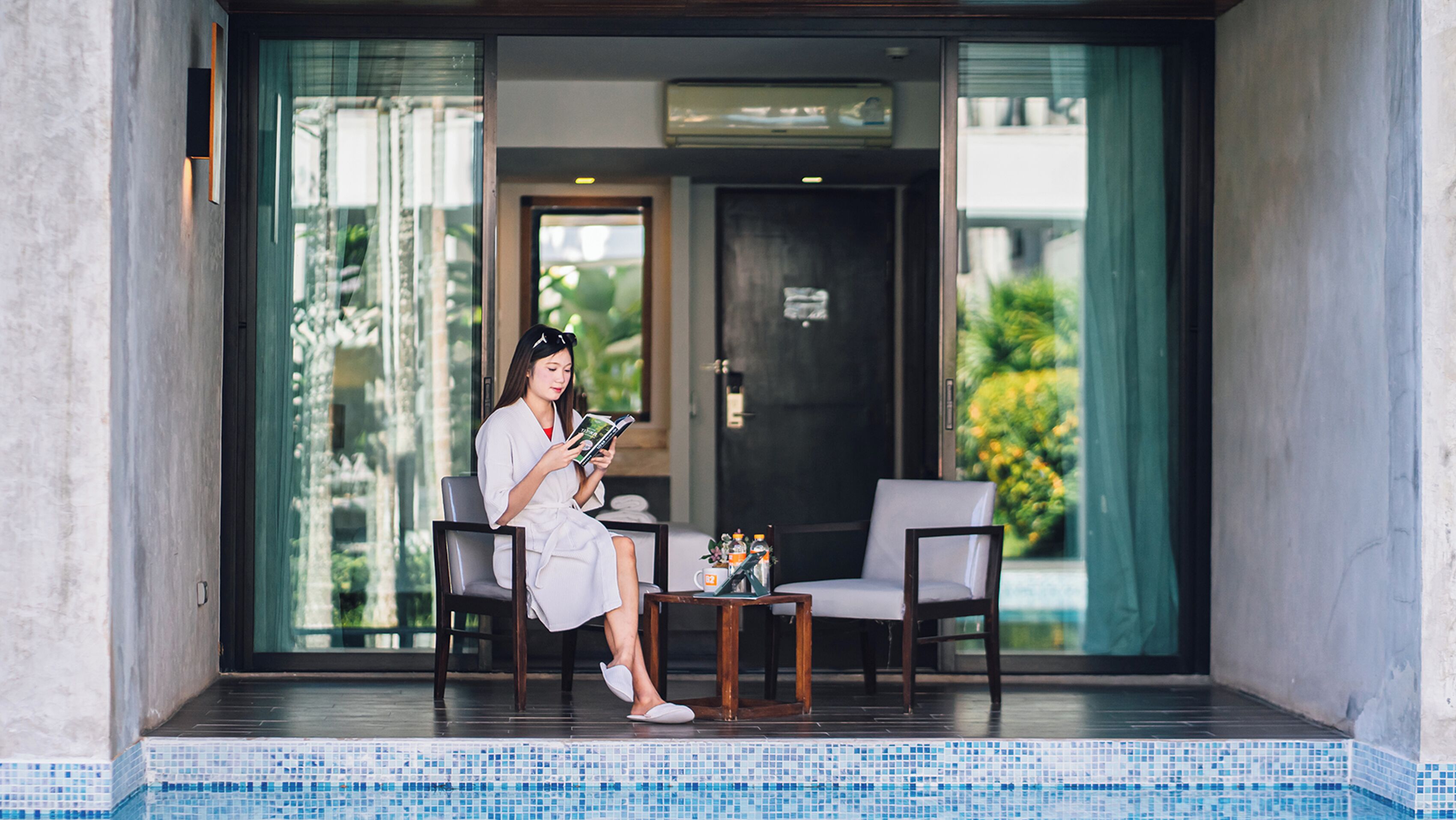 A woman sitting by a poolside chair reading a magazine, with a small table and a bottle of water in front of her.