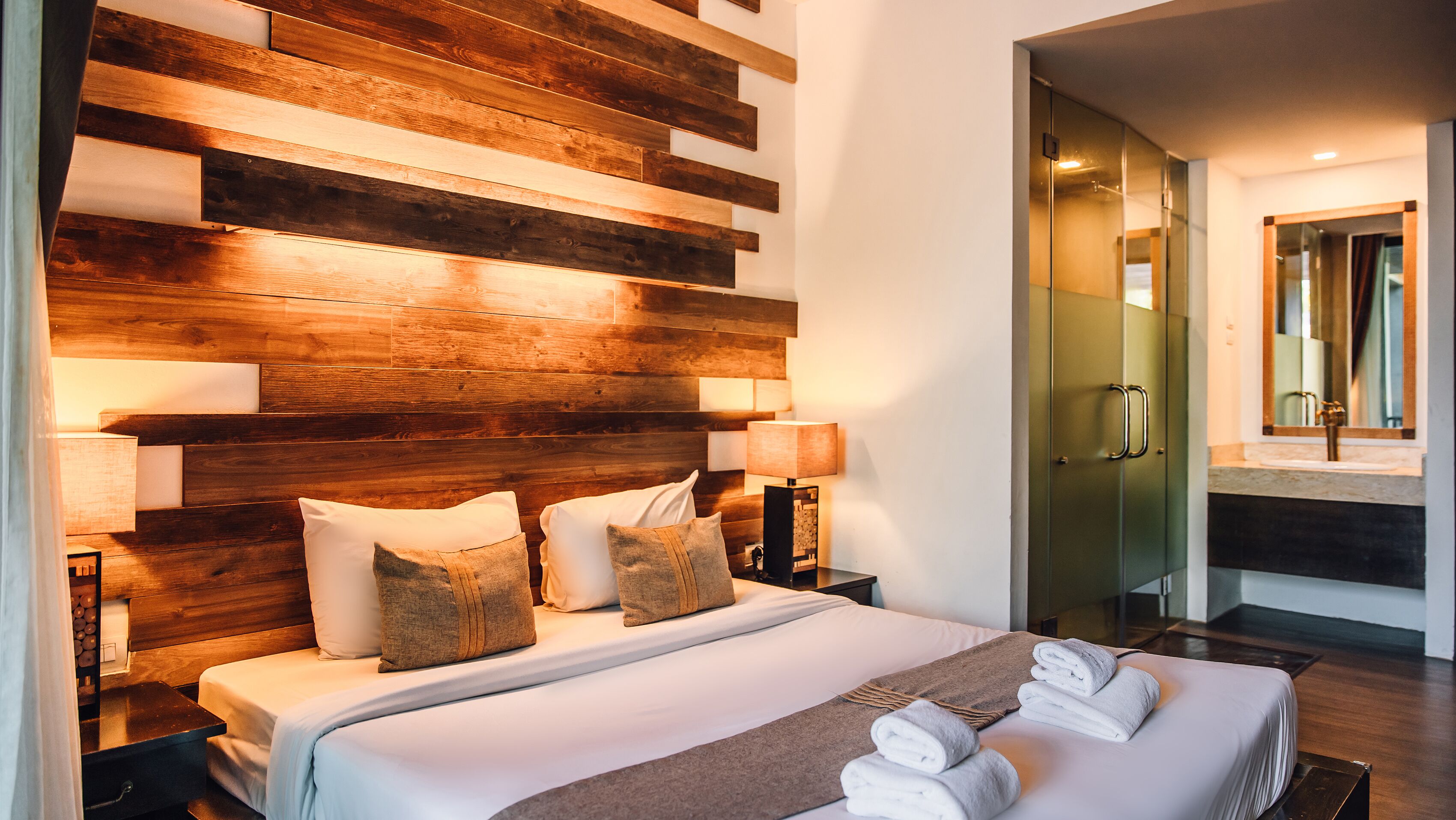 Une chambre d’hôtel moderne avec une tête de lit en bois, un grand lit avec du linge de maison blanc, deux lampes de chevet et une salle de bain visible à travers une porte en verre.