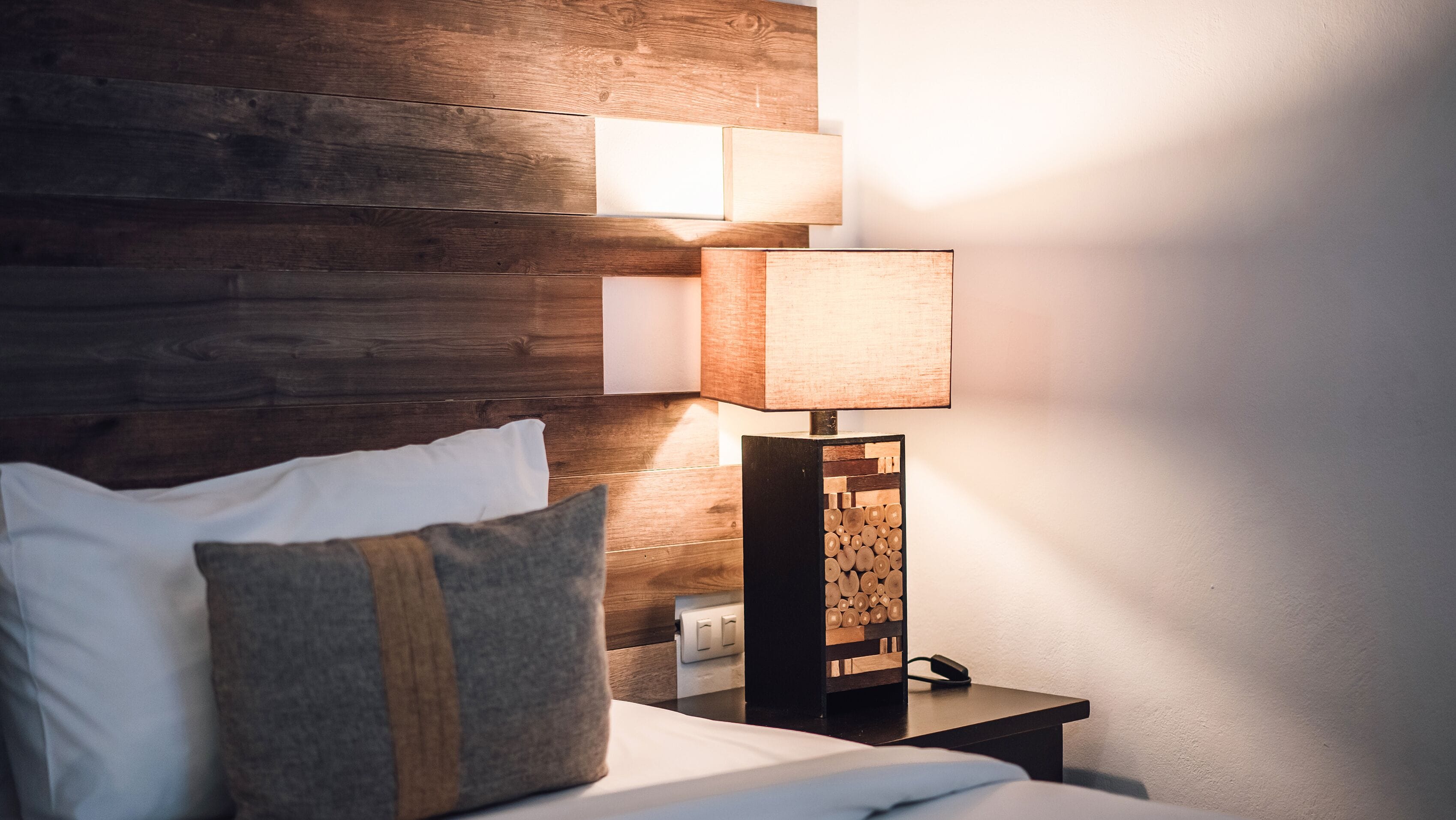 Una cama bien tendida con sábanas blancas y un almohadón gris, una mesita de noche con lámpara y un cabecero de madera.