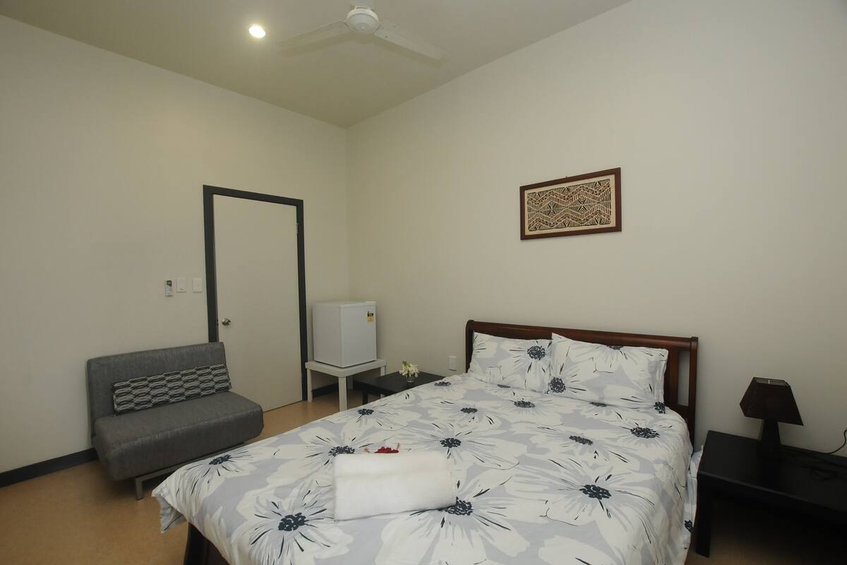 A bedroom with a bed, a sofa, a small table, a nightstand, a ceiling fan, and a wall-mounted artwork.