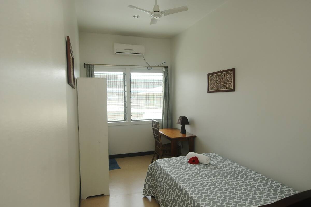 A small, clean room with a bed, a desk, a chair, a ceiling fan, and a window with curtains.