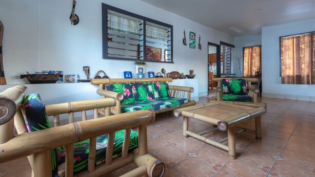 A room with a bamboo sofa set, a dining area with a table, and a kitchen in the background. - Talofa Inn (Apia)