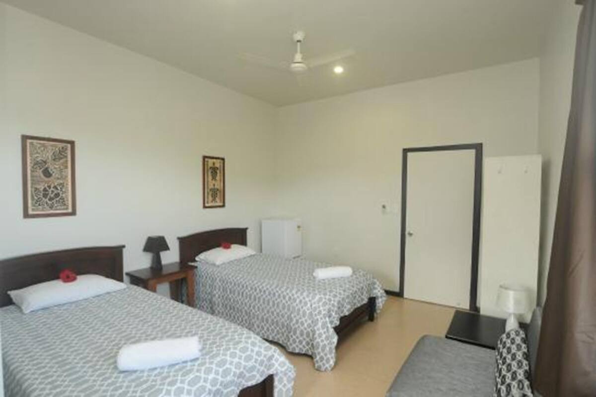 A hotel room with two beds, a desk, a chair, a wardrobe, and a ceiling fan.
