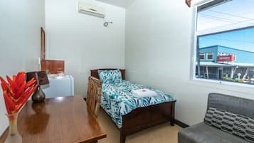 A small, tidy room with a bed, a wooden nightstand, a small table with a vase of flowers, a window with a view of a building, and a wall-mounted air conditioner.