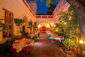 An outdoor dining area with tables and chairs, surrounded by plants and trees, and a building with arches in the background. - Hotel De L'Orient Pondicherry (Puducherry)