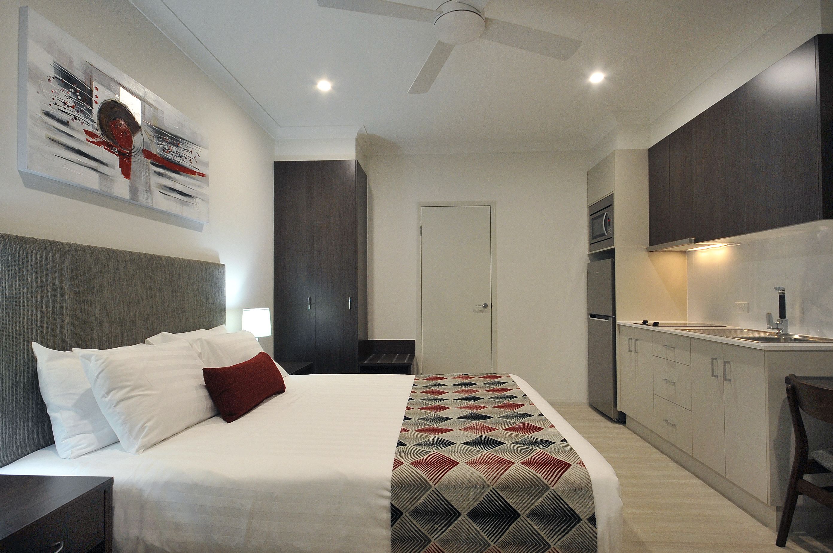 A modern hotel room with a large bed, a desk, a chair, a wardrobe, and a kitchenette.