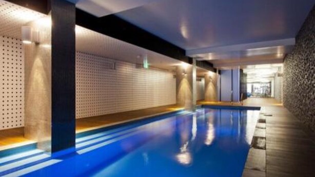 A modern indoor swimming pool with a blue floor, surrounded by walls with a perforated pattern and a stone-textured wall on one side. - StayCentral - NGV Arts Centre Retreat (Southbank)
