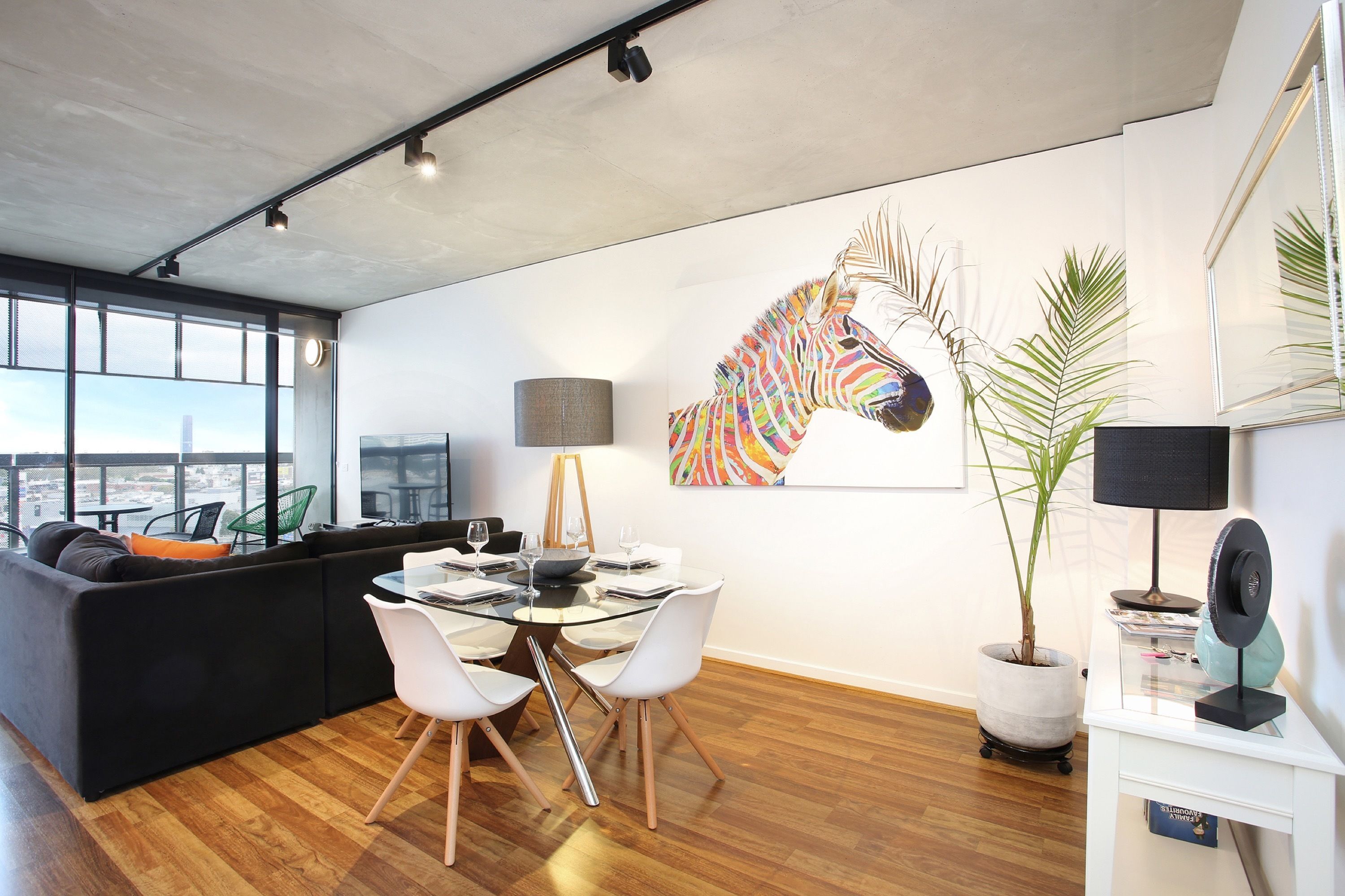A modern living room with a glass dining table, white chairs, a black sofa, and a large zebra mural on the wall.